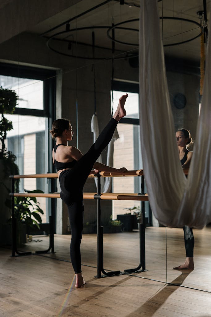 A woman practices barre exercises in a yoga studio with mirrors and hanging fabric.