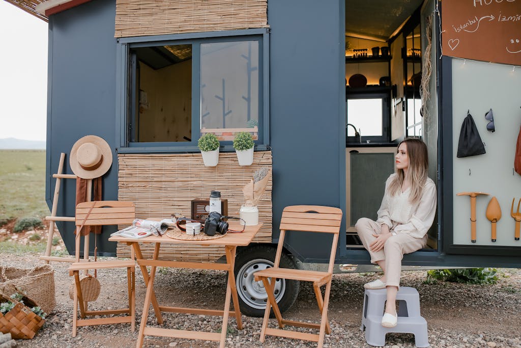 A woman sits outside a stylish tiny mobile home set in a natural landscape, enjoying a peaceful outdoor moment.