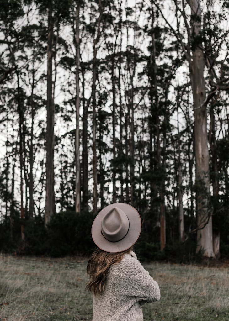 A woman stands thoughtfully in a hat, gazing at a peaceful, dense forest.