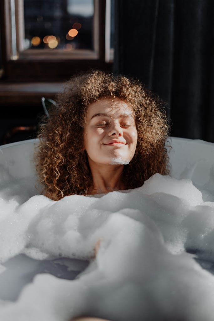 A young woman enjoys a relaxing bubble bath with natural light enhancing her curly hair.