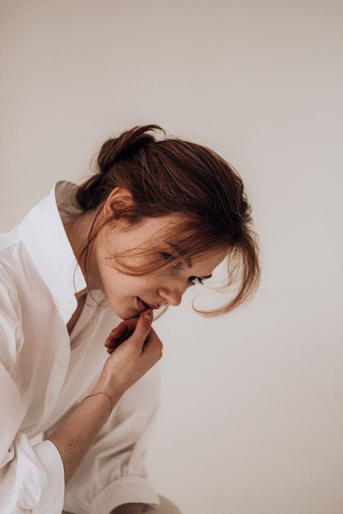 A young woman with dark hair pensively touching her chin, wearing a white shirt against a neutral background.