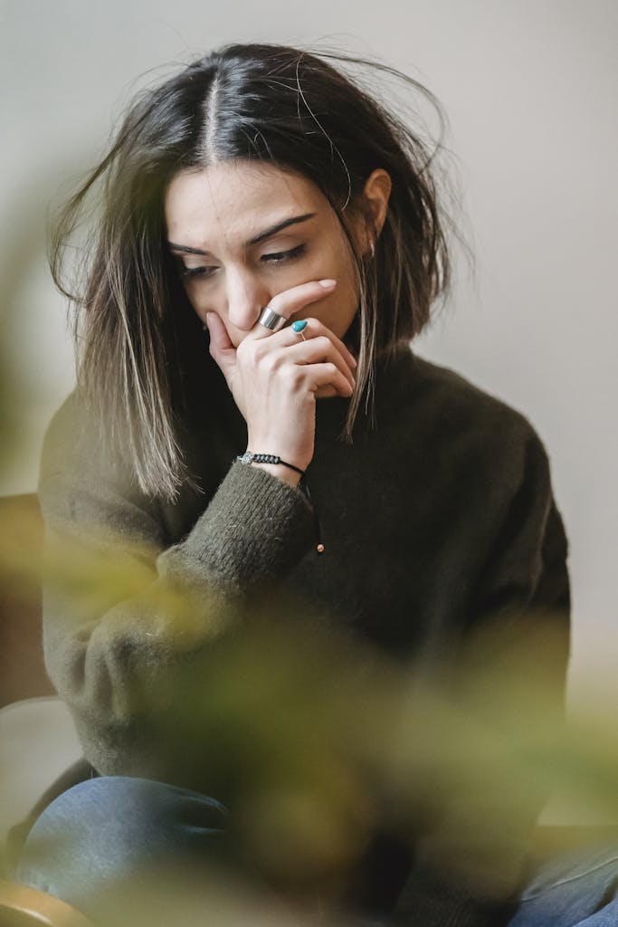 A young woman with short dark hair sits indoors, appearing deep in thought and contemplation.