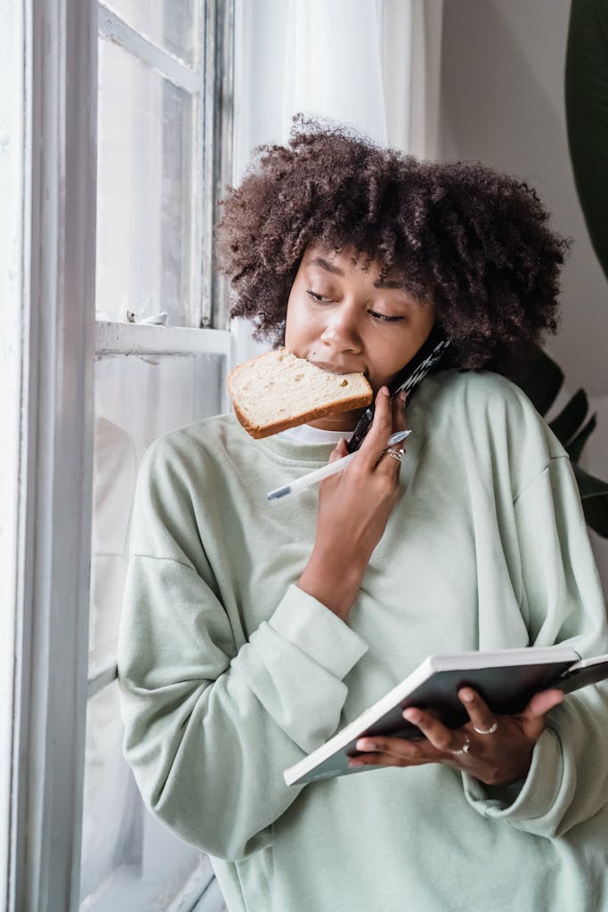 African American woman multitasks by a window with a sandwich and notebook, merging work and snack time.