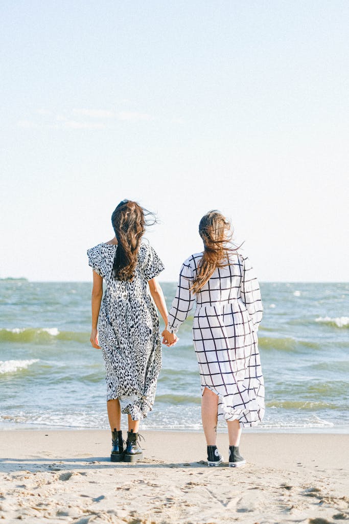 Back view of close women in dresses holding hands while standing on sandy beach near sea