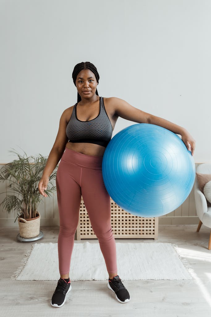 Black woman in activewear holding a large blue exercise ball indoors.