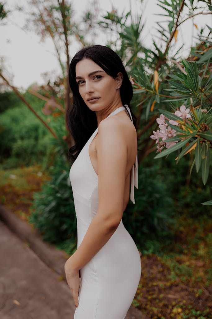 Elegant woman in white dress poses in lush outdoor setting with flowers.