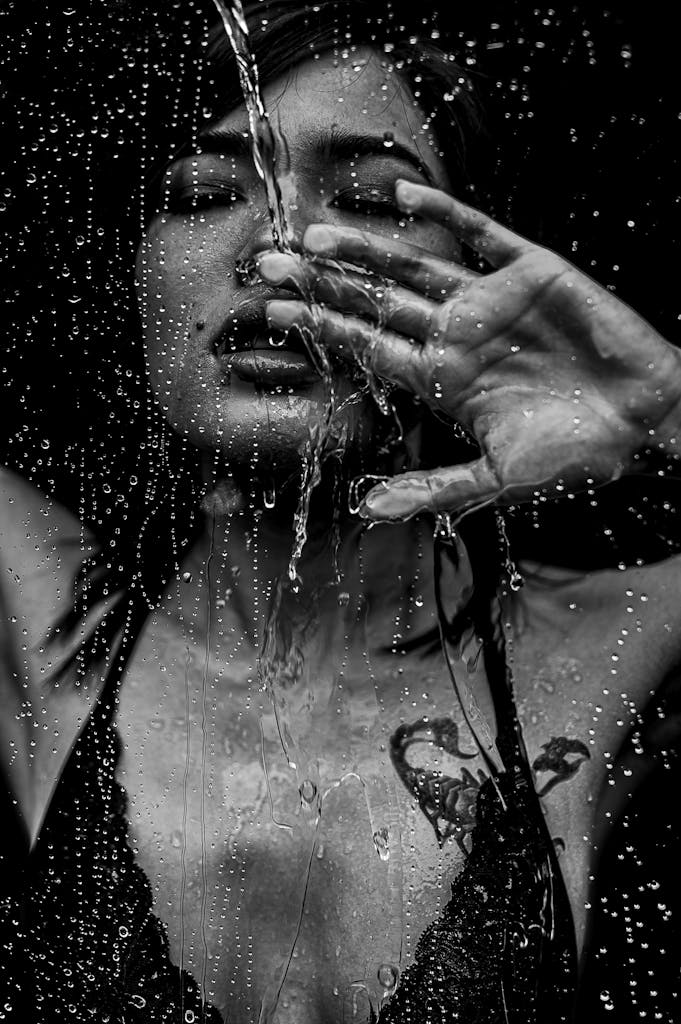 Emotive black and white portrait of a woman with water cascading down, highlighting tattoos.