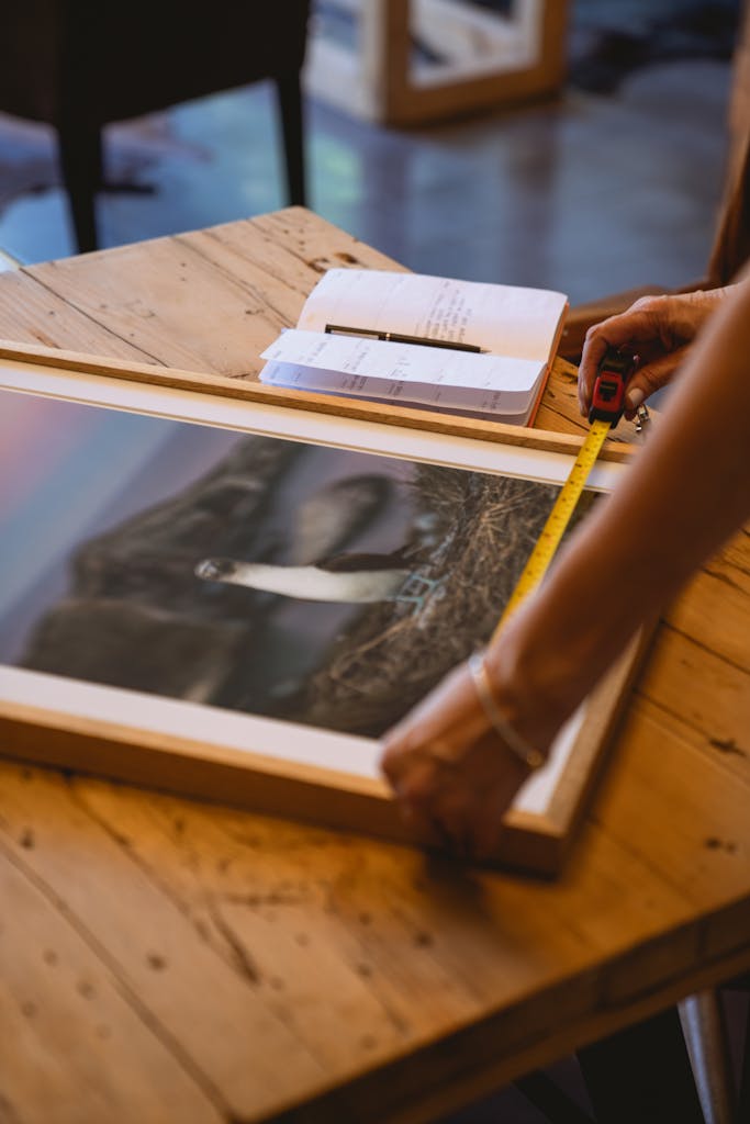 Person measuring a framed photograph with a tape measure on a wooden table indoors.
