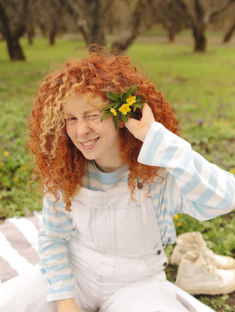Red-haired woman winking playfully with wildflowers in a lush green park.