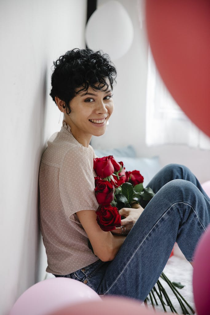 Smiling woman with short hair holding red roses, sitting indoors, embracing a cheerful and serene vibe.