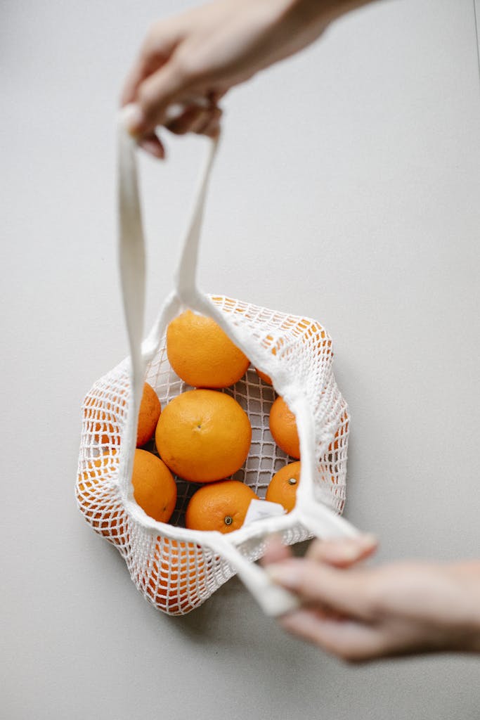 Top view of crop anonymous person with string bag full of ripe whole oranges placed on white background in room