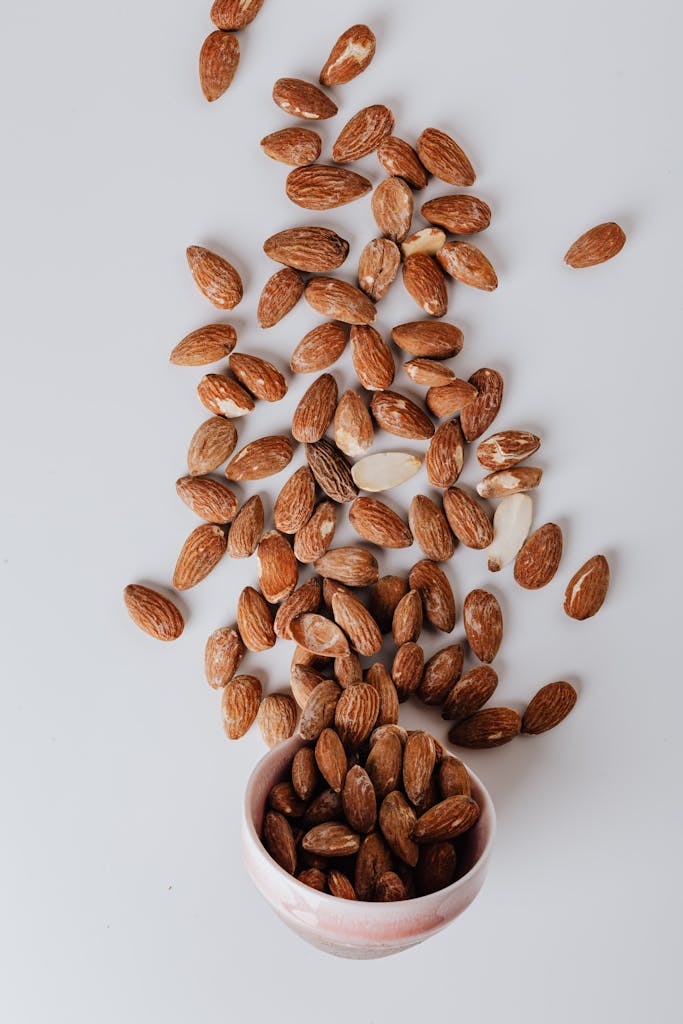 Top view of scattered raw almonds around a pink ceramic bowl on a clean white surface.