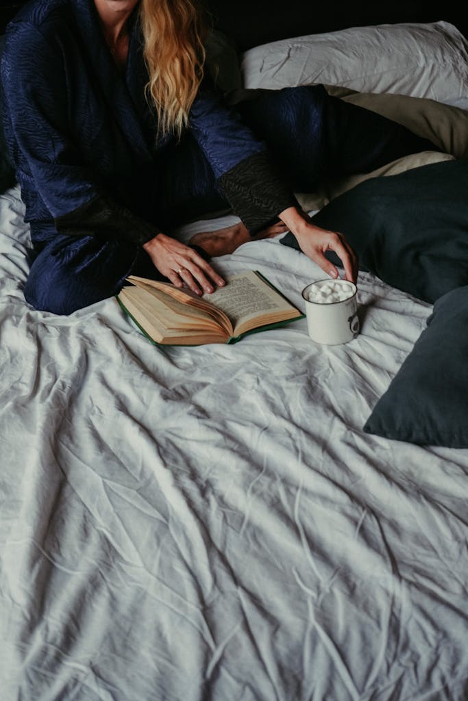 Woman in a robe enjoying a book and hot chocolate on a bed, creating a cozy indoor atmosphere.