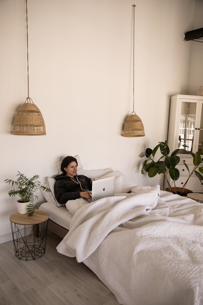 Woman relaxing in a cozy bedroom with a laptop, surrounded by plants and ambient lighting.