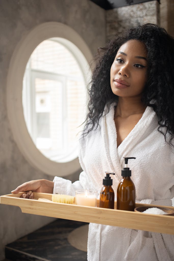 Young ethnic female in white bathrobe holding wooden tray with skin care products standing in bathroom and looking away