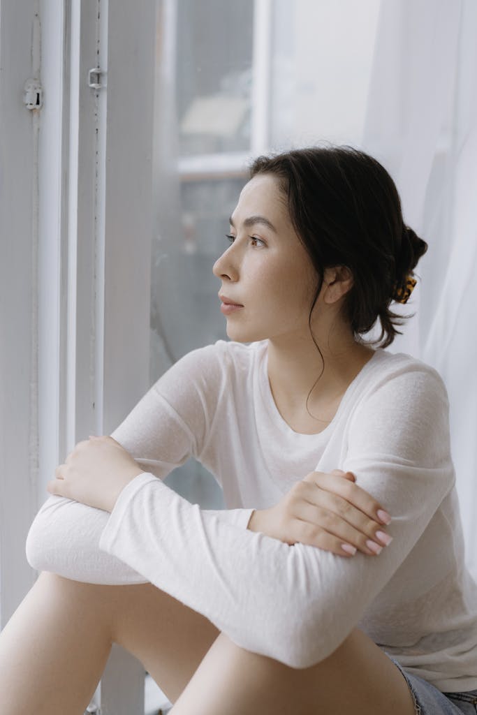 Young woman in white long-sleeve shirt thoughtfully gazing out a window indoors.