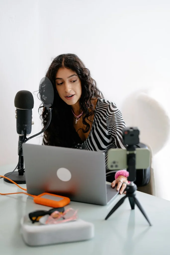 A young woman using a microphone and laptop to record a podcast indoors, showcasing a modern content creation setup.