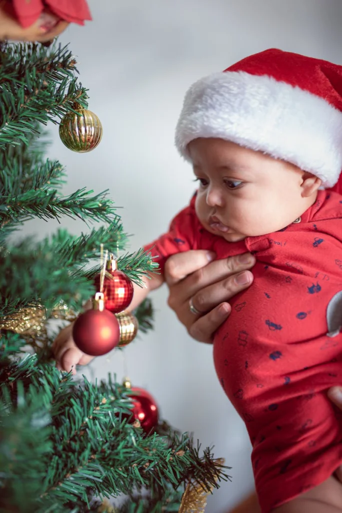 A cute baby in a Santa hat reaches for ornaments on a Christmas tree indoors.