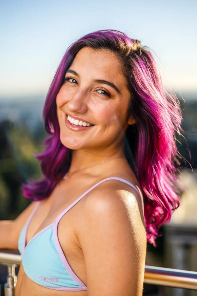 Smiling woman with vibrant purple hair enjoying a sunny day outdoors.