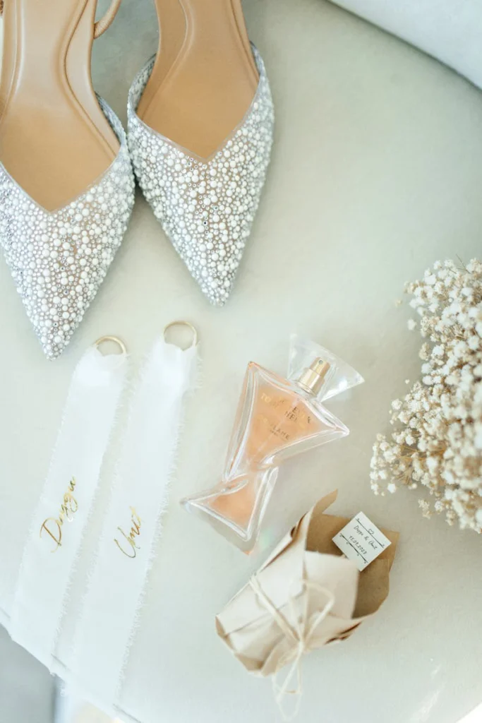 A flat lay of bridal accessories including elegant shoes, perfume, rings with ribbons, and flowers.