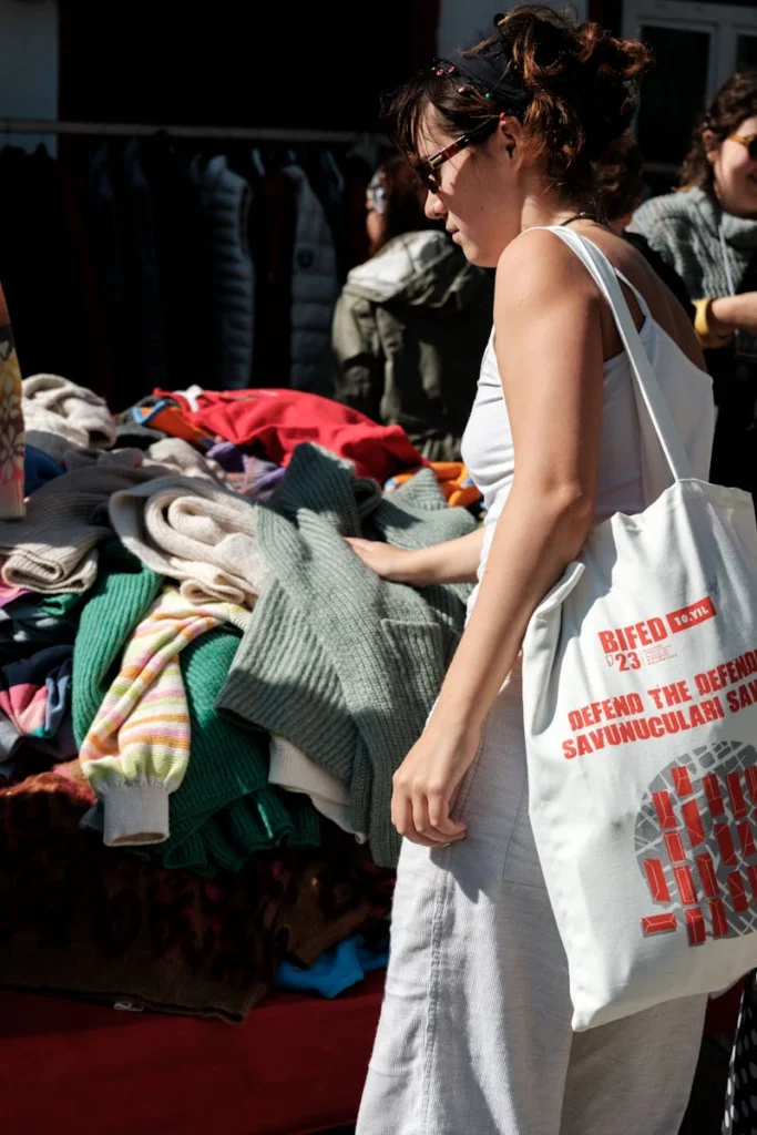 Woman browses clothes at an outdoor market, wearing sunglasses and a tote bag.