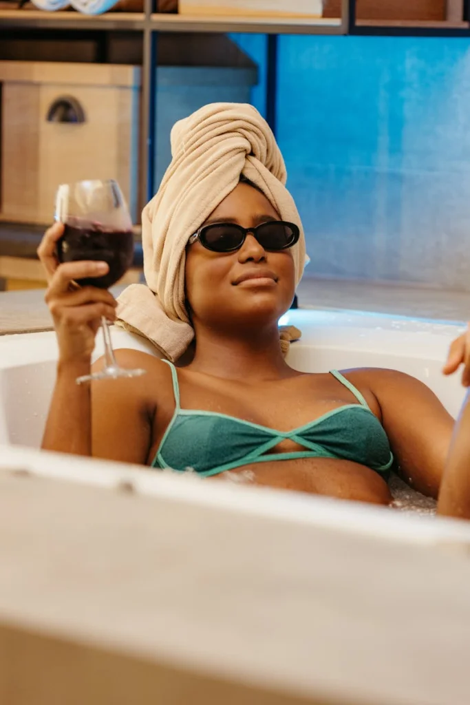 Woman in bikini and sunglasses enjoying a glass of red wine in a hot tub indoors.
