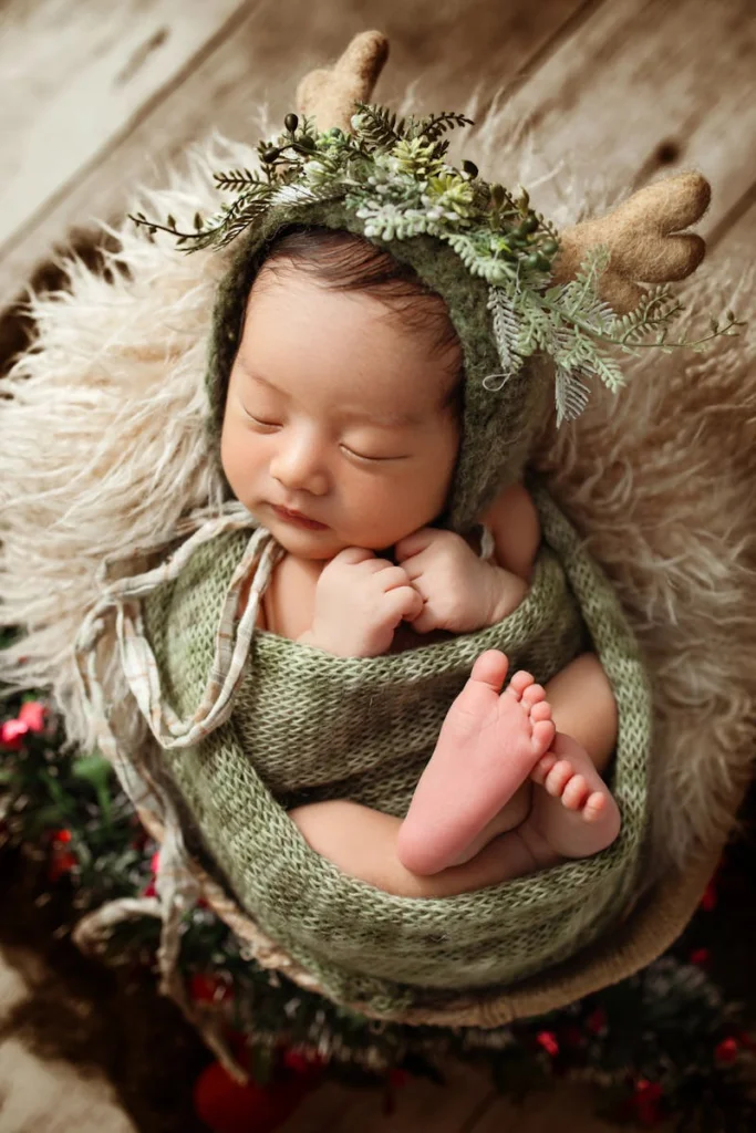 A cute newborn baby wrapped in green, adorned with an antler headband, sleeping peacefully in a cozy setting.