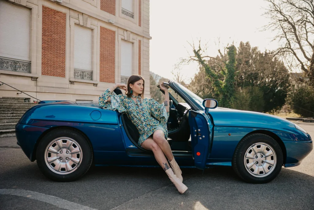 Young woman in floral dress poses by vintage blue convertible in sunny outdoor setting.