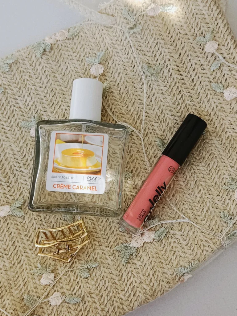 Elegant flat lay of Crème Caramel perfume, lip gloss, and hair clip on woven background.