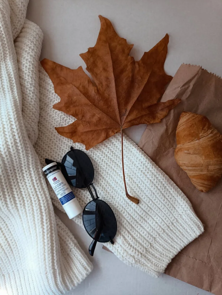 Cozy autumn flat lay featuring a sweater, sunglasses, leaf, and croissant.