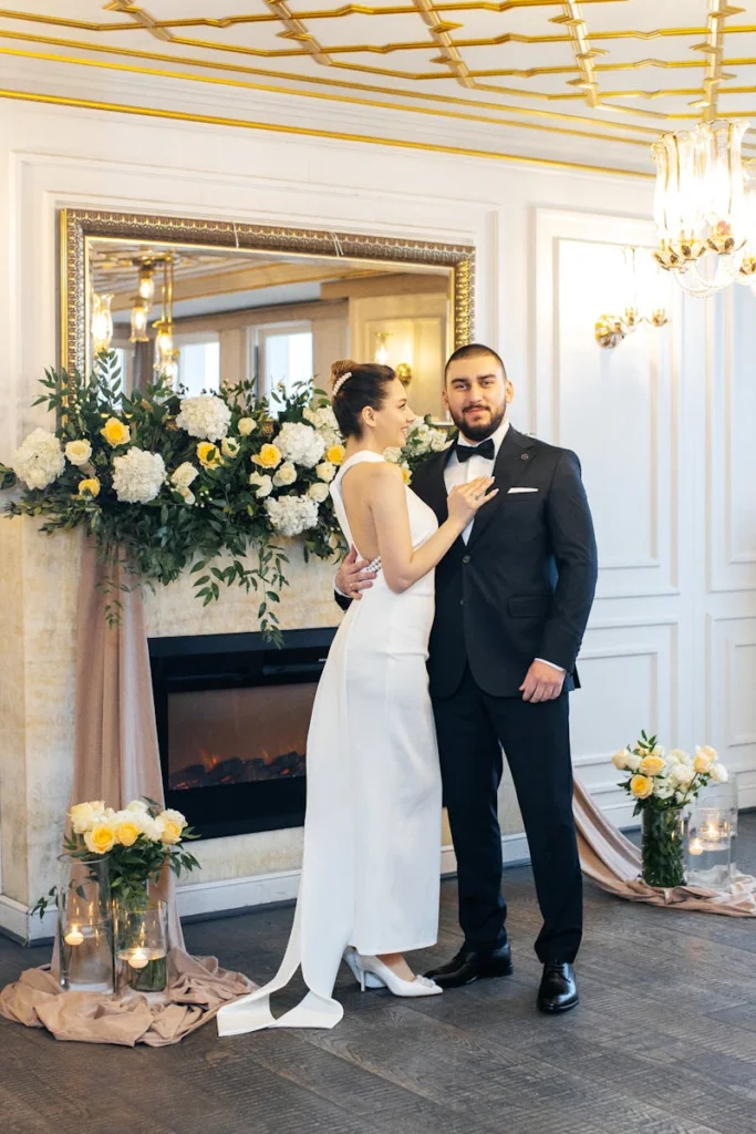 A stylish couple poses elegantly beside a decorated fireplace indoors.