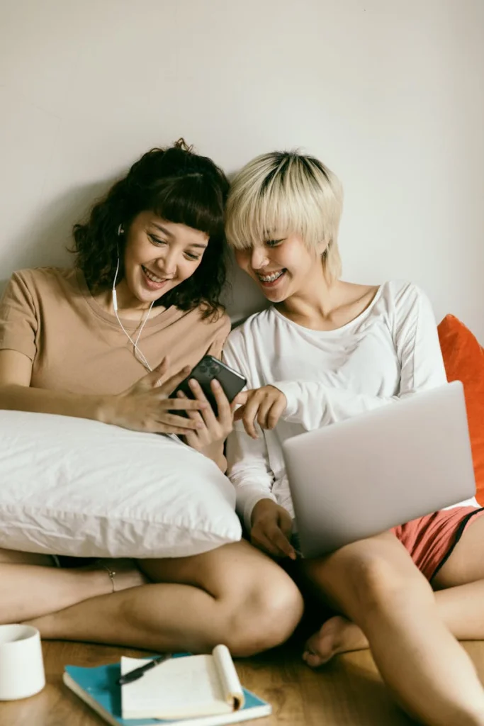 Happy smiling young Asian girlfriends in casual clothes sitting on floor with laptop and earphones and browsing smartphone together at home