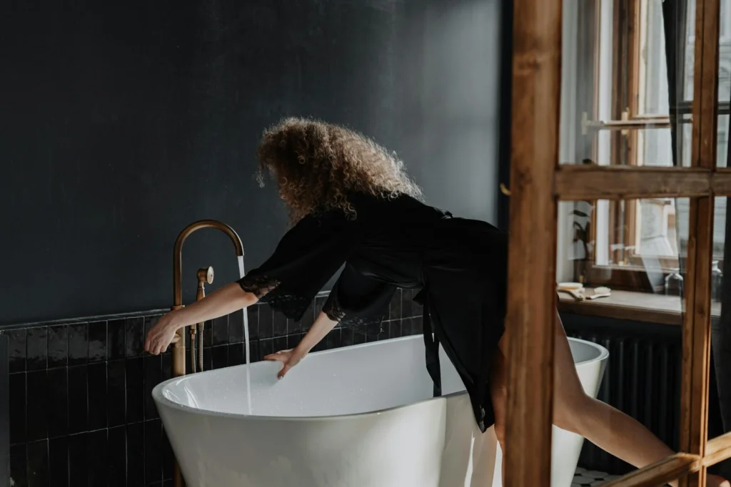 A young woman with curly hair prepares a bath in a stylish, modern bathroom with dark tile and natural light.