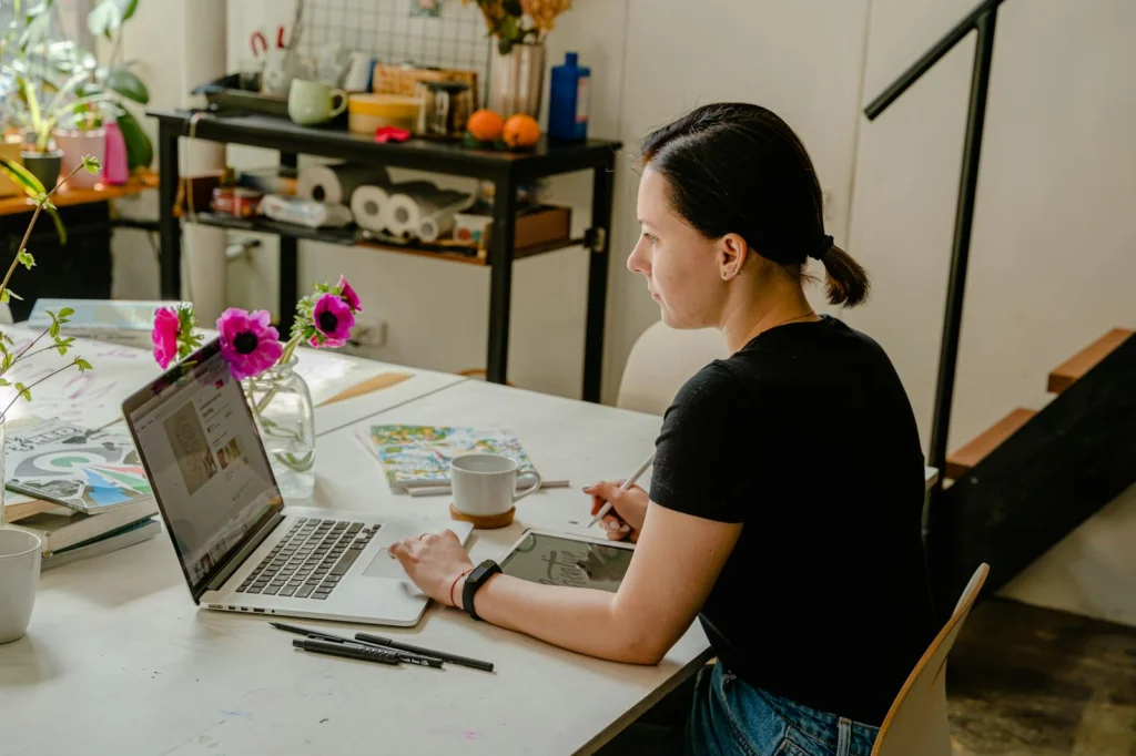 Woman working from home with a laptop and tablet in a cozy workspace surrounded by plants and decor.