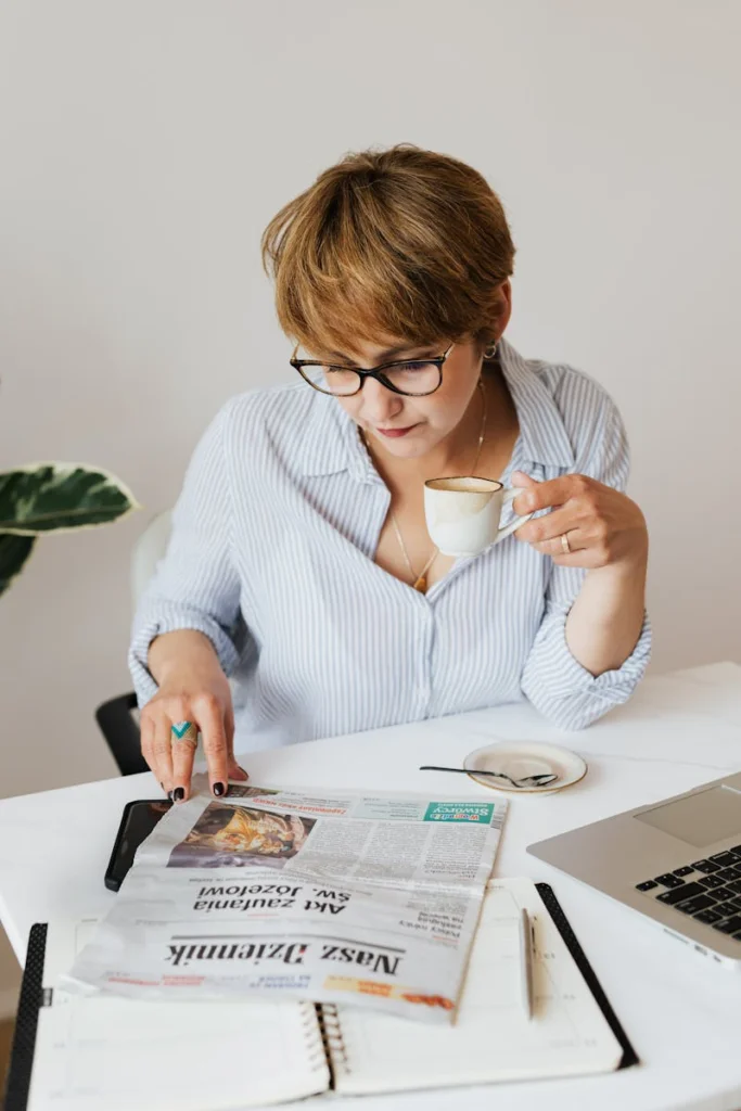 Thoughtful female in light blue shirt with eyeglasses sitting at desk with notebook and laptop while reading newspaper and enjoying cup of fresh coffee