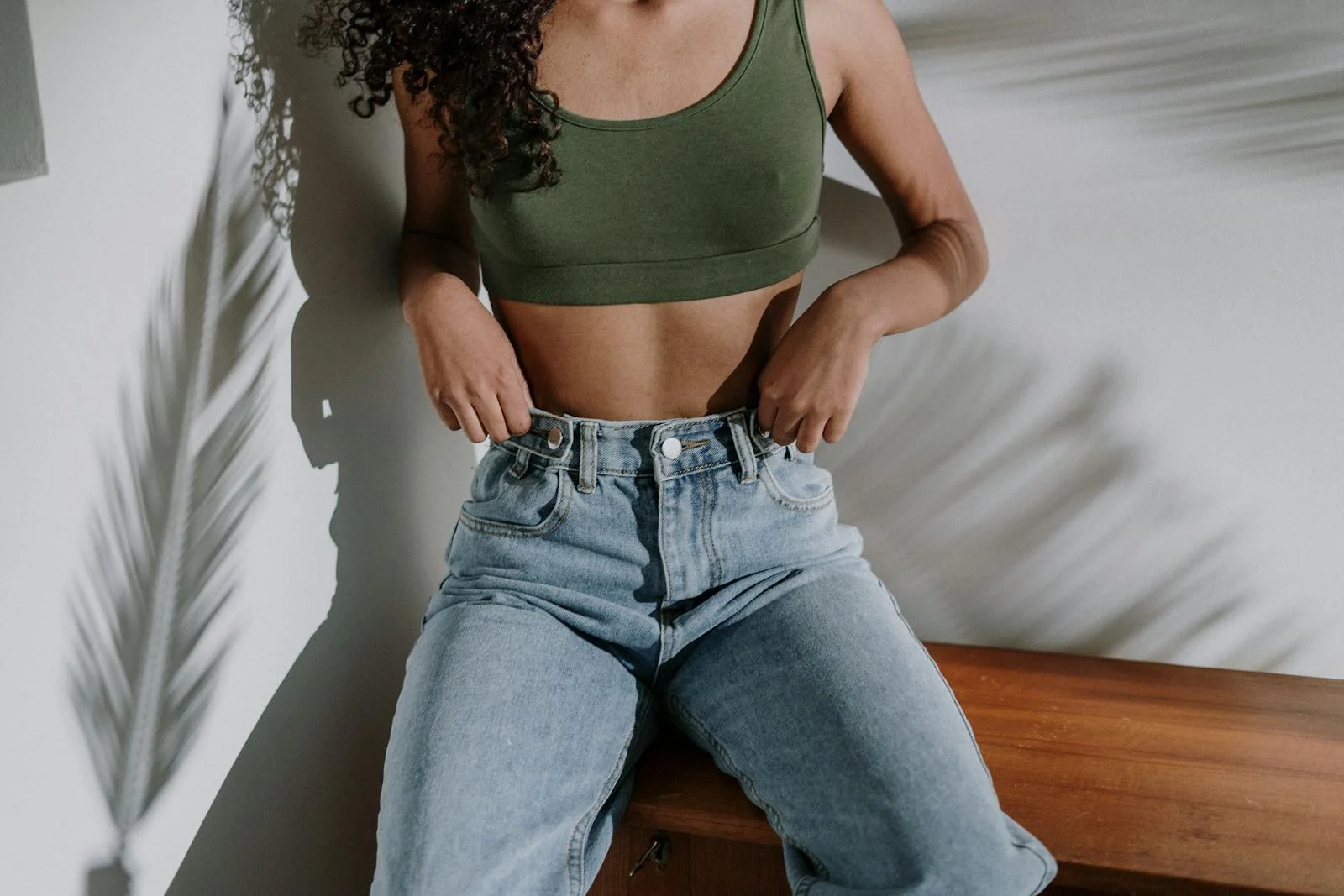 A woman in jeans and a crop top sits calmly, with tropical shadows on the wall.