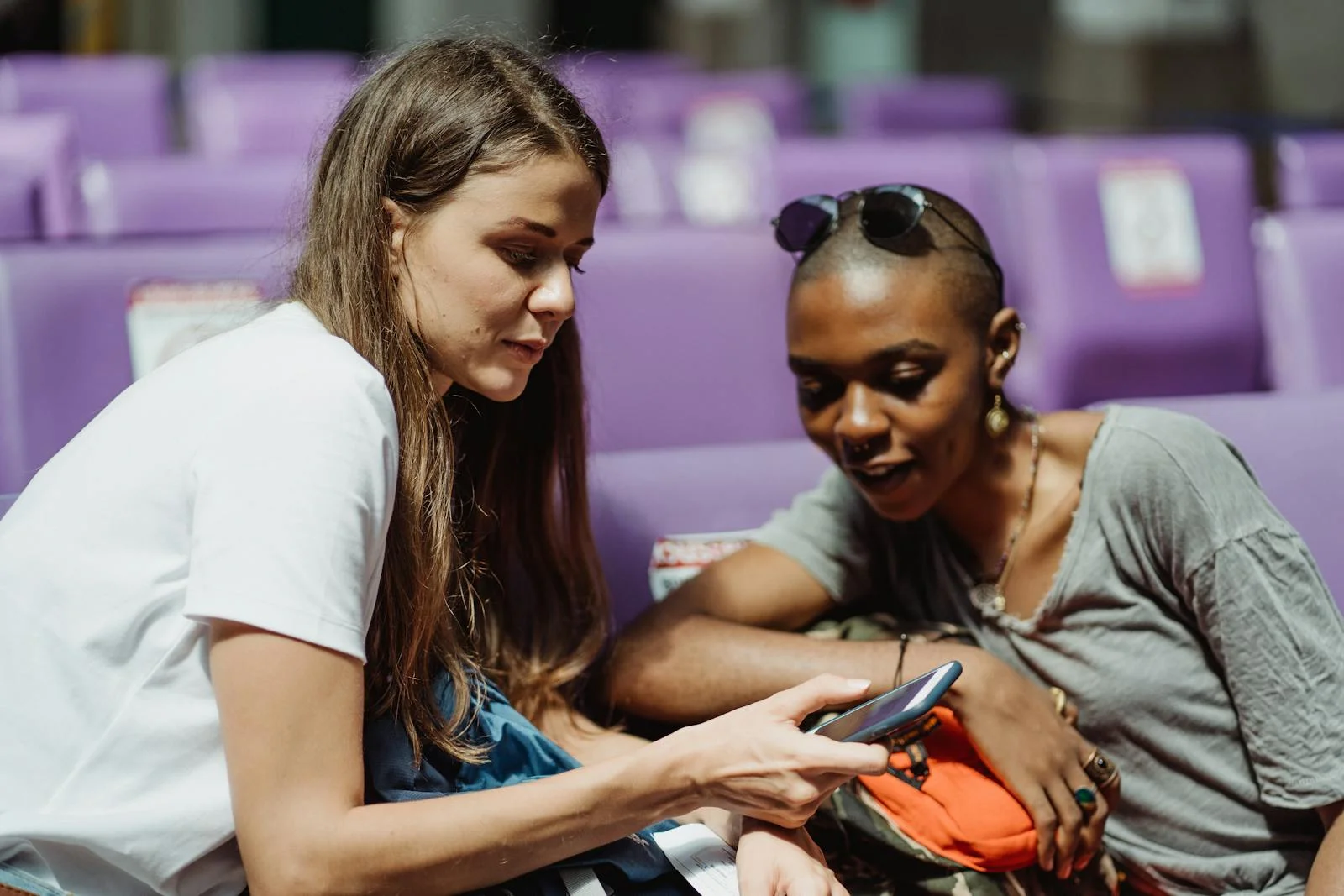 Two adult women sitting together and sharing a smartphone in a public waiting area with purple seats.