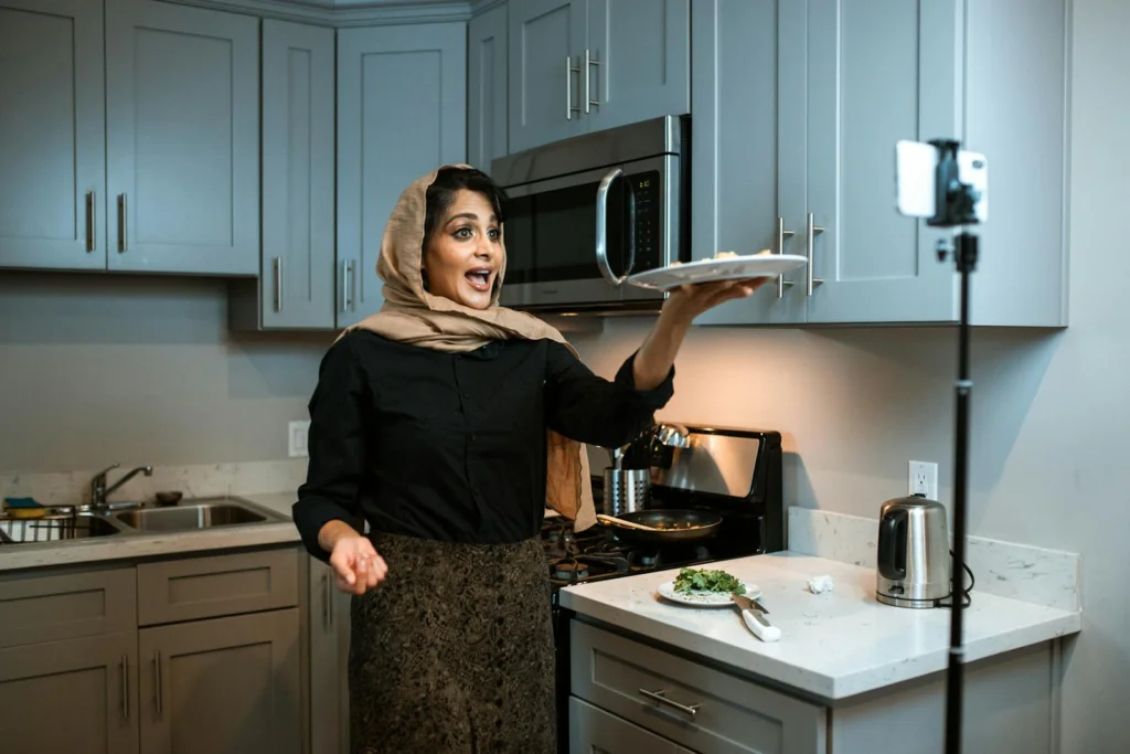 Smiling woman in hijab recording a cooking vlog in her kitchen.