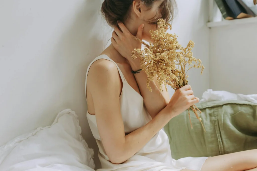 A woman in a white tank top holding dried flowers, sitting comfortably indoors.