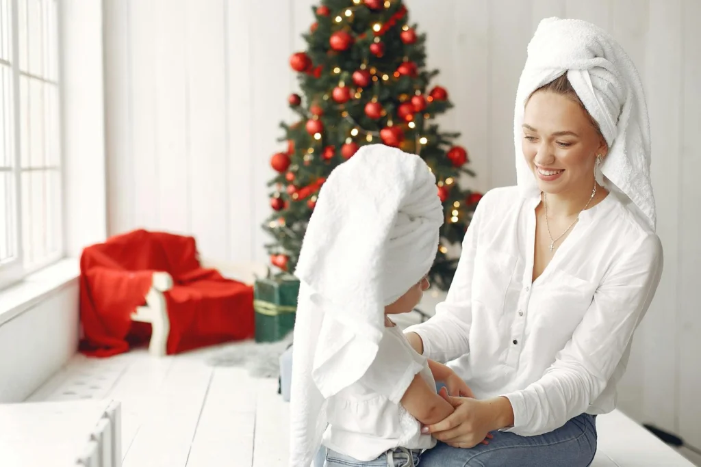 A mother and daughter sit happily with towels on heads next to a decorated Christmas tree.