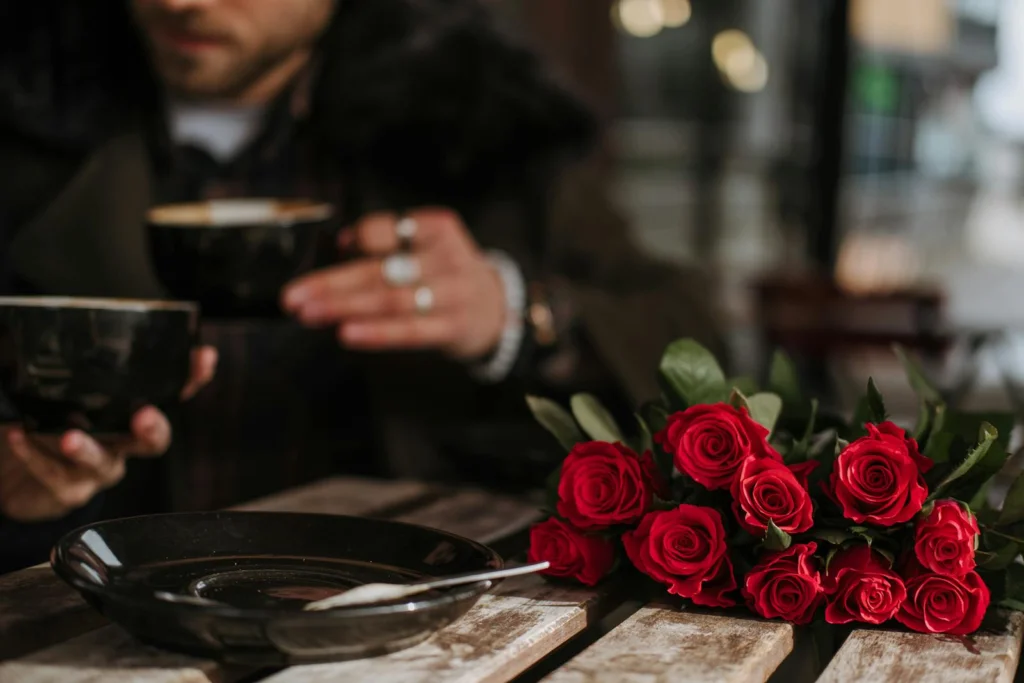 Crop unrecognizable man and woman in stylish outfit drinking coffee in street cafe sitting at table with bunch of elegant red roses