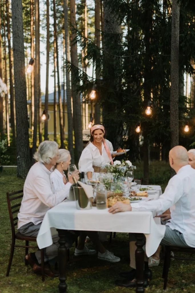 Senior adults enjoying a lively dinner party outdoors in a forest setting with decorative lighting.