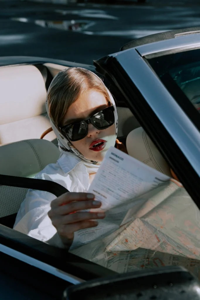 Elegant woman in a convertible reading a map, showcasing classic fashion style on a sunny day.