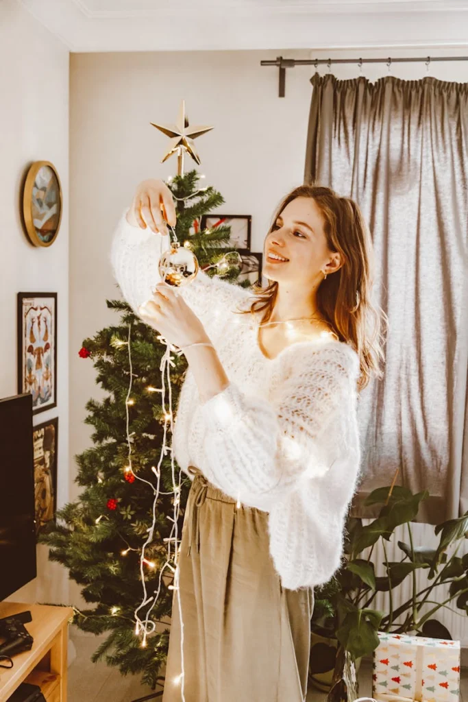 Woman decorates Christmas tree with a bauble at home, creating a festive atmosphere.