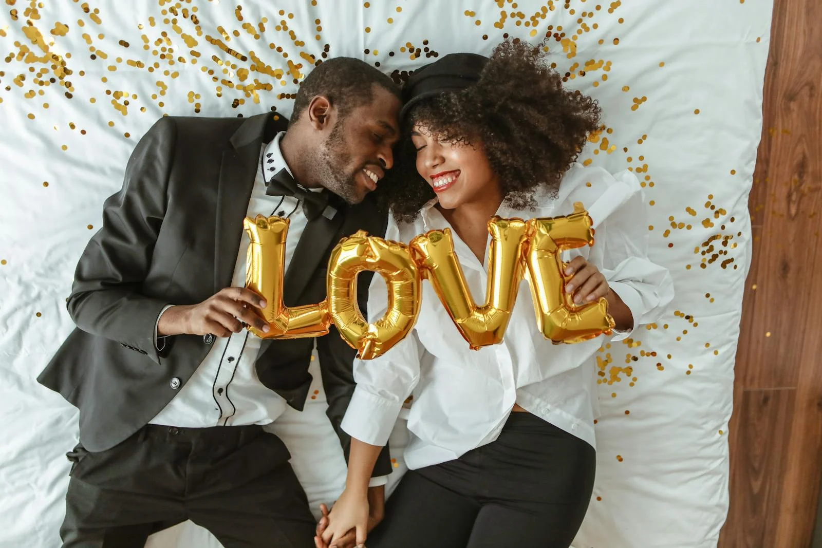 Smiling black couple lying on bed with LOVE balloons, joyful and intimate moment.