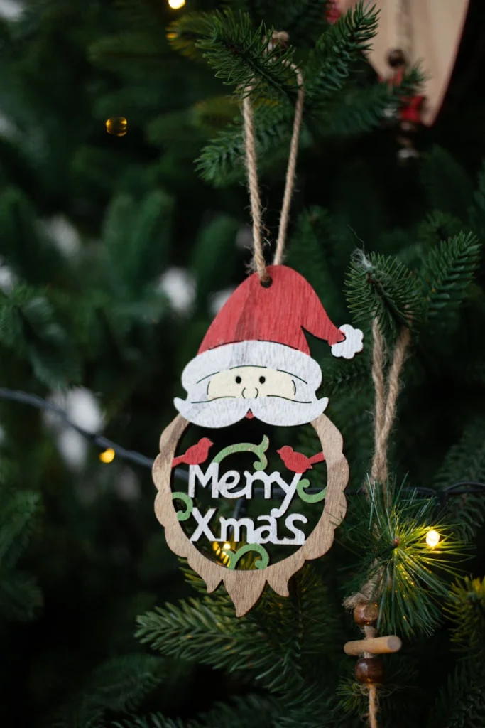 A close-up of a wooden Merry Xmas ornament hanging on a fir tree with lights.
