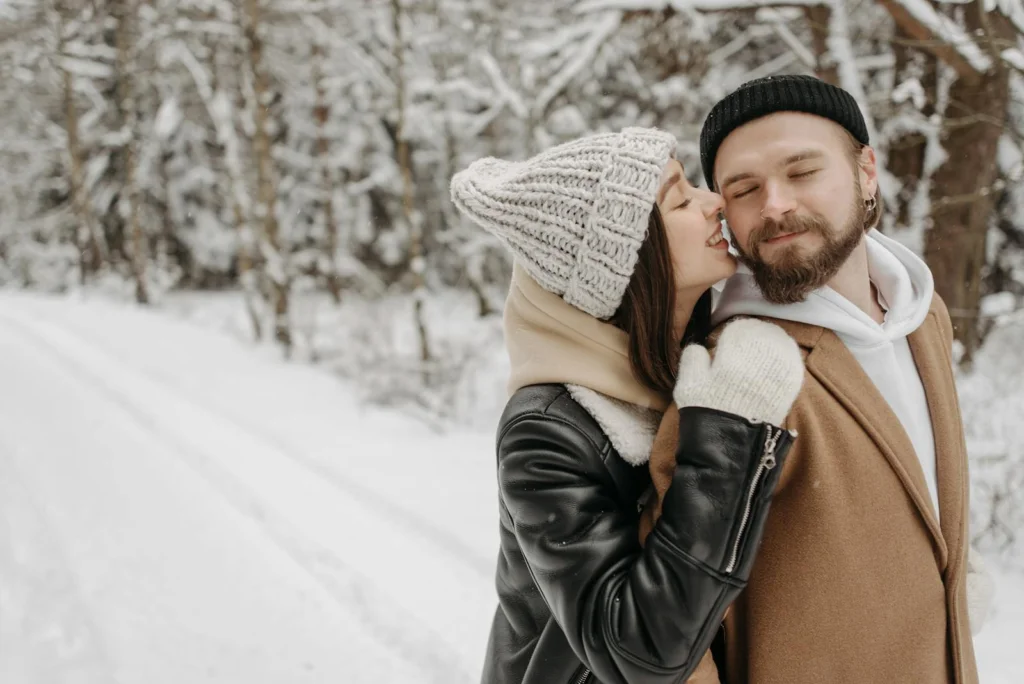 A loving couple embraces in snowy winter attire surrounded by a serene snowy forest.