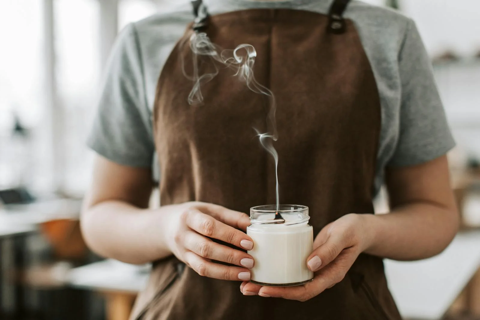 Close-up of a person in an apron holding an unlit candle with smoke in a workshop setting.