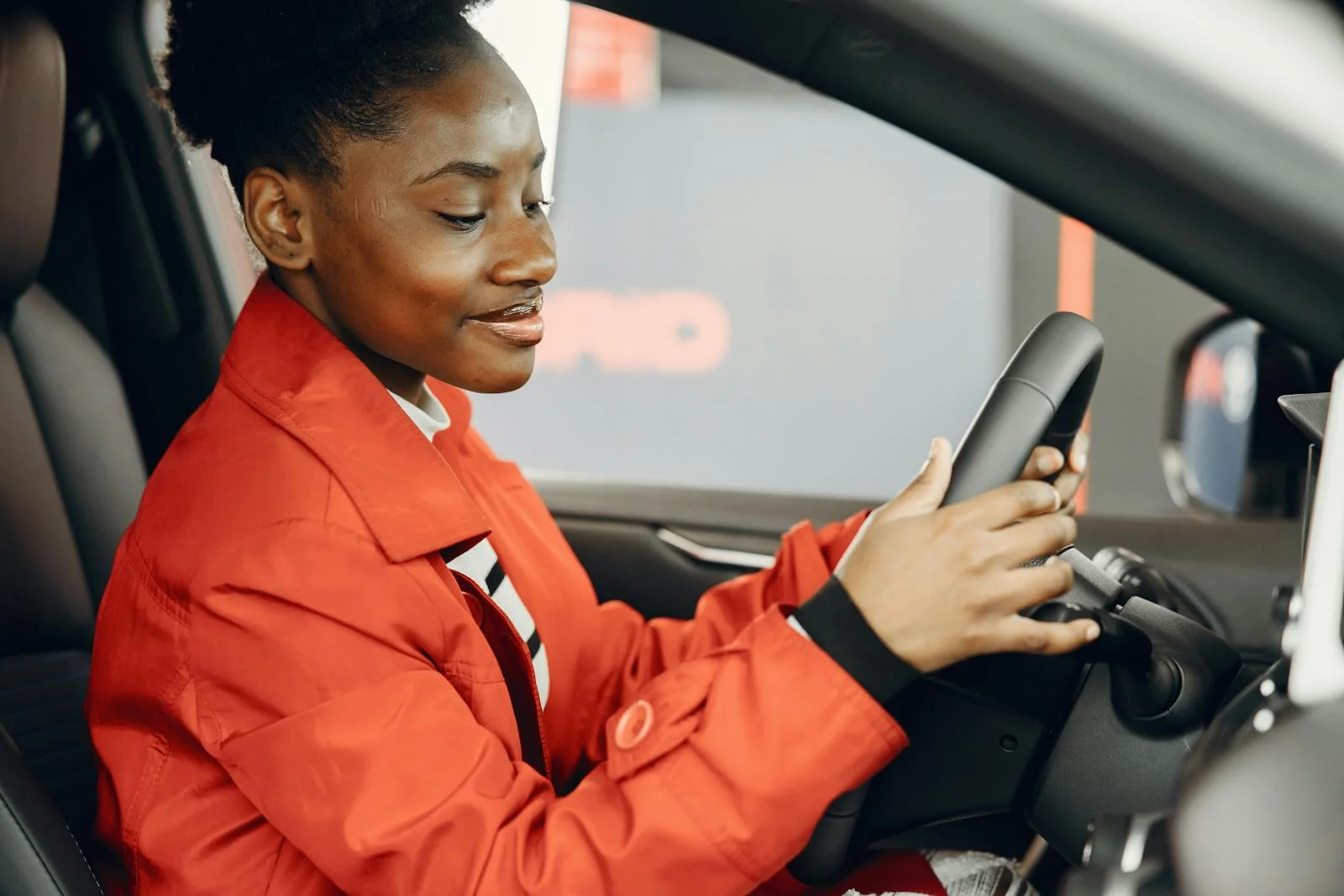 Black woman in orange jacket sitting in a car holding the steering wheel.