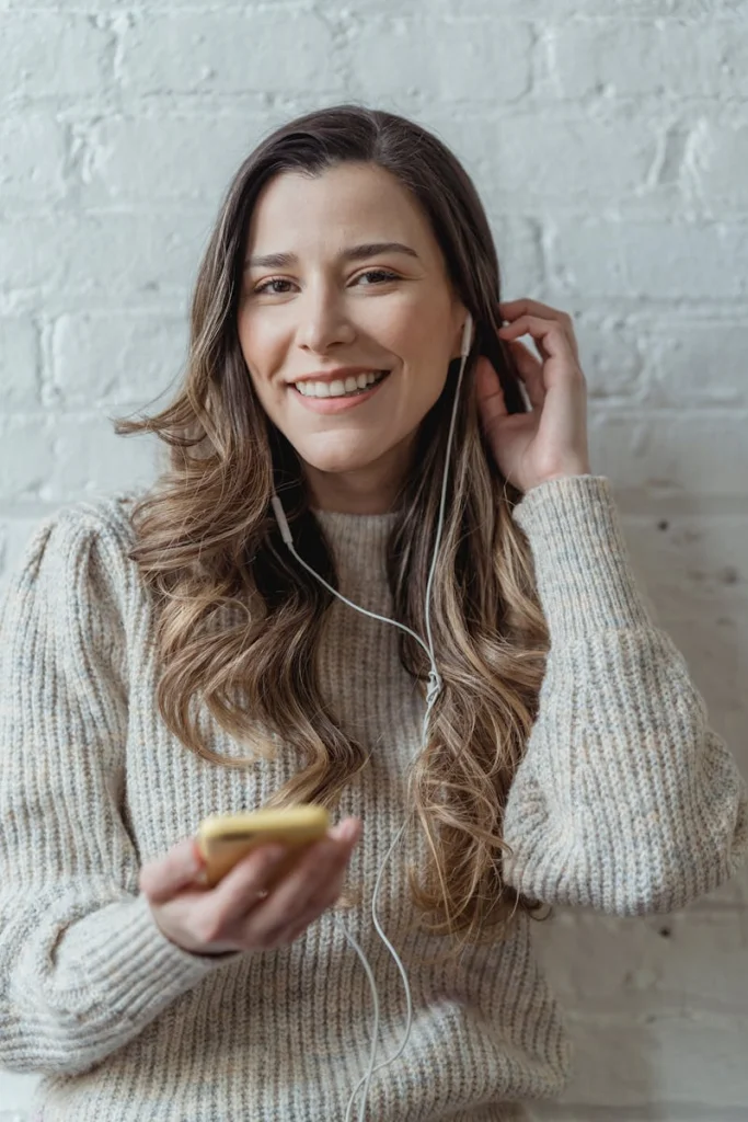 Young content female in sweater and earphones browsing mobile phone and looking at camera while standing against brick wall and touching hair